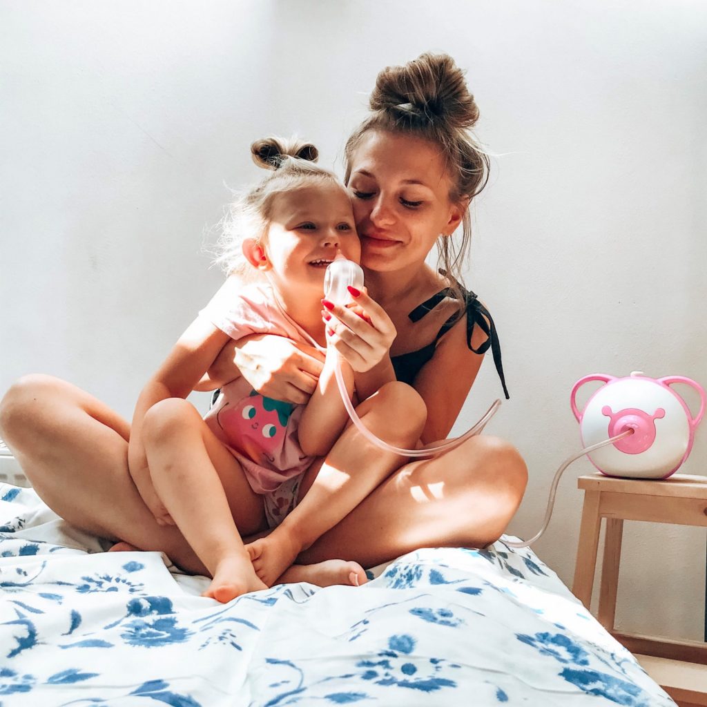 Smiling mom and her daughter sitting on a bed and using Nosiboo Pro electric nasal aspirator